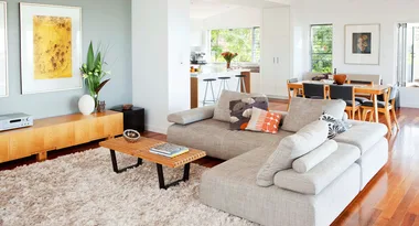 Modern living room with a grey sectional sofa, wooden coffee table, and dining area in the background.