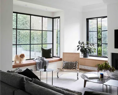 Bright living room with large windows, grey sofa, armchair, round coffee table, and a small dog on a window seat.