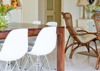 Dining room with a wooden table, white chairs, a wicker chair, a vase of orange tulips, and a lamp on a sideboard.