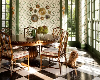 Elegant dining room with green lattice ceiling, floral wallpaper, round table, bamboo chairs, and a small dog on checkered floor