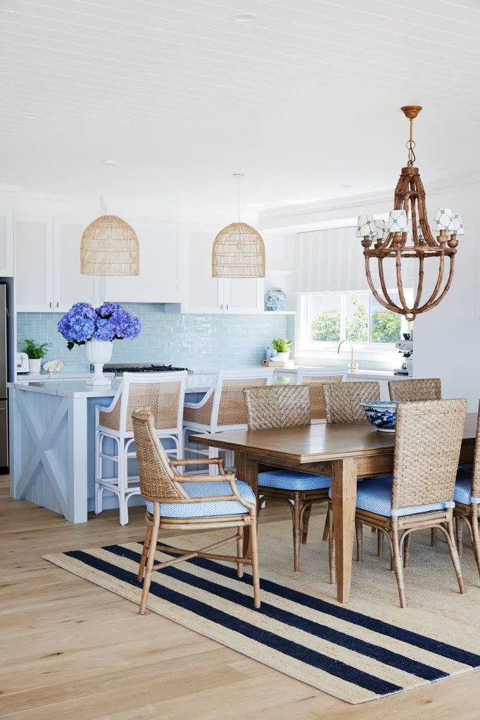 Bright kitchen with wicker chairs, a wooden table, blue splashback, rattan pendant lights, and a striped rug. The blue splashback matches the blue kitchen island and the blue upholstery on the seat cushions of the dining table.