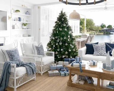 Coastal living room with a Christmas tree, white furniture, blue accents, gifts beneath tree, large windows, and chandelier.