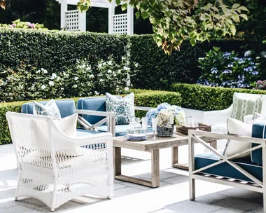 Outdoor patio with white and blue furniture, surrounded by greenery and flowers, under a tree canopy.
