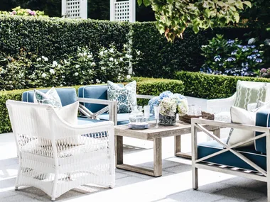 Outdoor patio with white and blue furniture, surrounded by greenery and flowers, under a tree canopy.