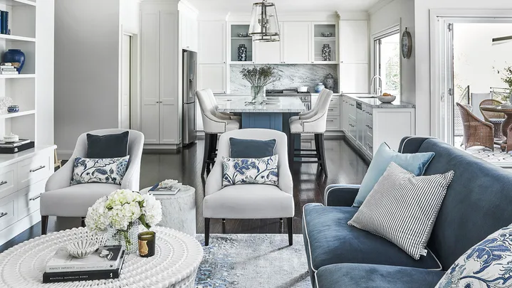 Elegant living room and kitchen with blue and white decor, featuring a sofa, armchairs, and a white wicker coffee table.