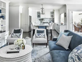 Elegant living room and kitchen with blue and white decor, featuring a sofa, armchairs, and a white wicker coffee table.