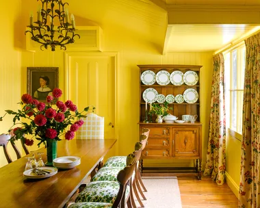 Yellow dining room with wooden table, floral chairs, chandelier, and hutch of plates. Vase with red flowers on table.