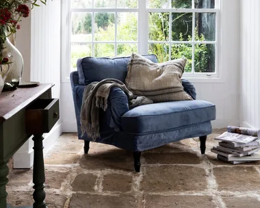 Cozy room with a blue armchair, cushions, and a blanket by a large window; plants and magazines nearby.