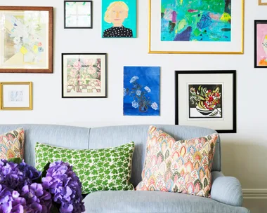 Colorful gallery wall above a light blue sofa with patterned pillows; vase of purple flowers on table in foreground.