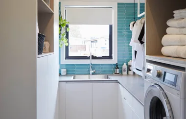 Laundry room with a window, teal tiles, sink, plants, washer, shelves with towels and detergent bottles.