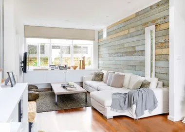 Modern living room with white sofas, rustic wood accent wall, large windows, and a coffee table with decor items.