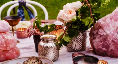 Outdoor table with pink crystals, flowers, metallic vases, and teacups set against a grassy background.