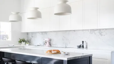 Bright modern kitchen with marble countertops, white cabinets, hanging lights, plants, and freshly baked bread on an island.