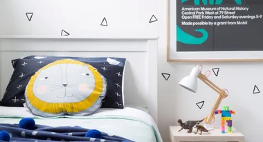 Cozy kids' room with lion face pillow, geometric decor, and a side table with lamp, toys, and framed museum poster.