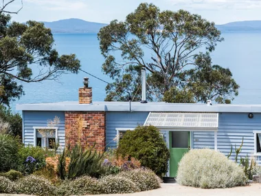 This fishing shack has been renovated into a colourful and pattern-filled Airbnb in Tasmania. The blue panelled exterior matches the blue of the sea views visible beyond the house.