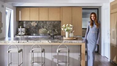 Bec Judd in the kitchen of her Arthurs Seat home, wearing a blue long sleeve top and matching long skirt with blue high heels. The kitchen has oak flooring, timber veneer joinery and marble benchtops.