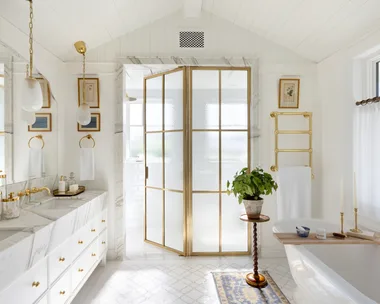 An all-white ensuite with gold glass doors, a freestanding bath and a marble vanity.
