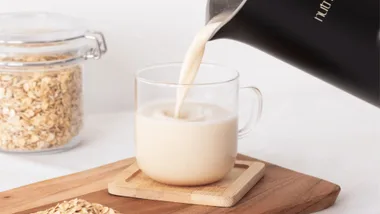 Pouring oat milk into a clear mug on a wooden tray, with a jar of oats in the background.