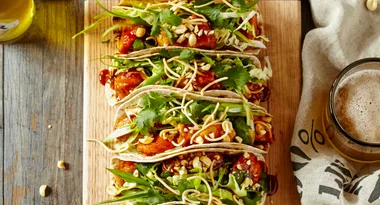 Tacos filled with crispy chicken, herbs, and nuts on a wooden board beside a glass of beer.