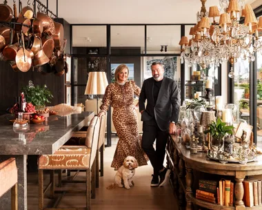 A stylish couple stands in a luxurious open plan kitchen and dining room with a small dog, surrounded by chandeliers and decor. The woman is Chyka Keebaugh, of Real Housewives of Melbourne fame and the man is her husband, Bruce Keebaugh.
