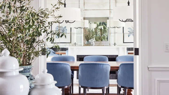Dining room with blue chairs, white chandeliers, plants, and art on walls, seen through a doorway.