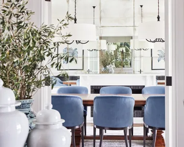 Dining room with blue chairs, white chandeliers, plants, and art on walls, seen through a doorway.