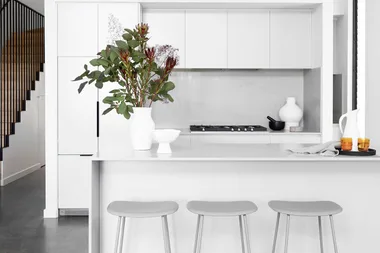 Modern white kitchen with a minimalist design, featuring bar stools, a countertop with flowers, and a staircase.