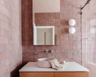A main bathroom featuring pink zellige wall tiles, white floor tiles, wooden joinery, a round basin, an arched mirror and two round wall sconces beside a shower. The tapware is brushed nickel in a curved style. The bathroom design is cohesive.