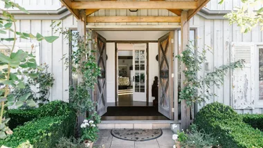 Rustic house entrance with wooden doors, green plants, and a mat on the stone path.