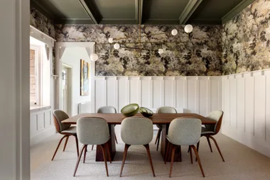 Dining room with a wooden table, eight chairs, modern lighting, and floral wallpaper.