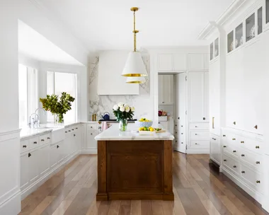 Bright kitchen with white cabinets, wooden floors, white pendant lights above a large kitchen island, which has marble countertop. A bowl of lemons is on it.