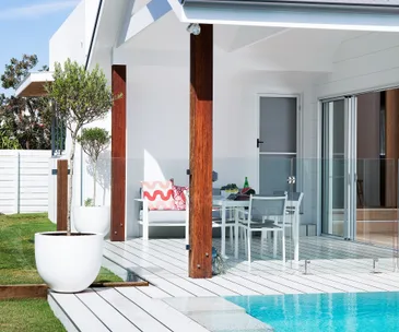 Modern patio with white furniture, glass railing, and pool, featuring wooden posts and a potted tree.