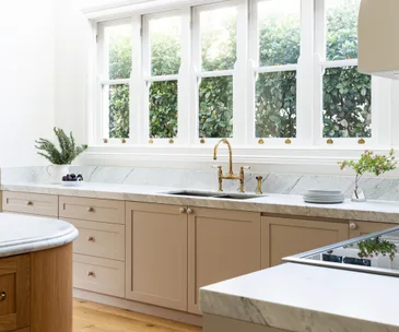 Stylish kitchen with marble countertops, beige cabinets, brass tap, large windows, and green plants outside.