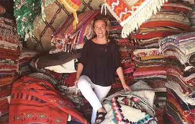 A woman smiling in a shop filled with stacks of colorful woven rugs and textiles.