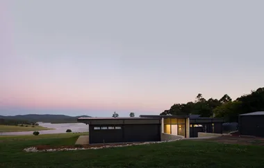Modern house with large windows at sunset, overlooking a serene lake and forested hills.