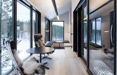 Interior of modern cabin with large windows, forest view, sleek furniture, and fur-covered chairs.