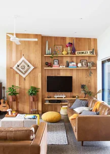 Cozy living room with leather sofa, wooden wall, mounted TV, shelves with decor, guitar, and plants, creating a warm ambiance.
