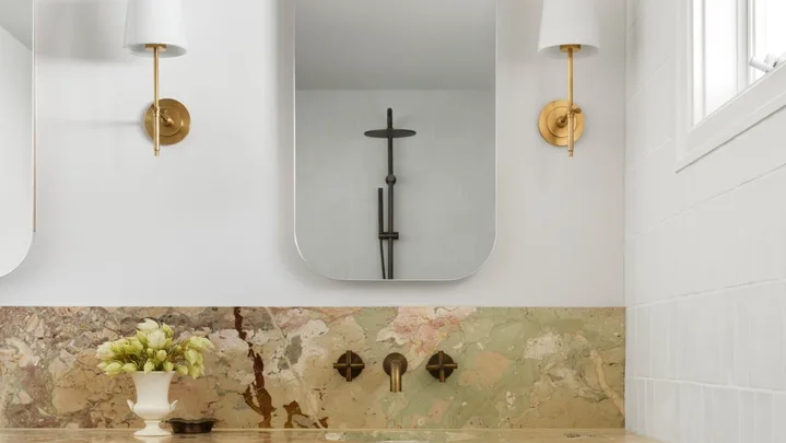 A bathroom vanity, with marble as the benchtop and splashback. The vanity has a deep apron, made from the same marble, which has warm tones complemented by antique brass tapware. Underneath the apron, oak joinery creates storage. Twin mirrors are positioned above twin basins in the vanity, with a wall sconce on either side.