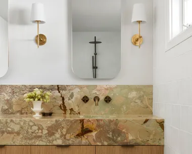 A bathroom vanity, with marble as the benchtop and splashback. The vanity has a deep apron, made from the same marble, which has warm tones complemented by antique brass tapware. Underneath the apron, oak joinery creates storage. Twin mirrors are positioned above twin basins in the vanity, with a wall sconce on either side.