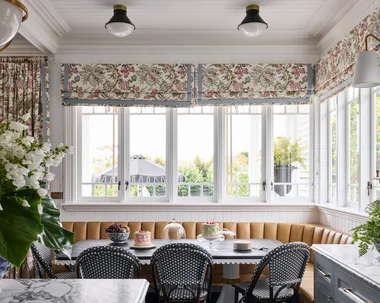 Elegant kitchen with a breakfast nook in front of the kitchen island. This is built around the corner of the room, underneath windows with floral Roman blinds. A dining table is surrounded on two sides by built-in banquette seating, with dining chairs on the other two sides. Marble countertops are visible in the foreground and the dining table is set with cakes and fruit.