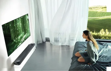 A woman watches a wall-mounted TV displaying a forest scene; sheer curtains and a scenic view are in the background.