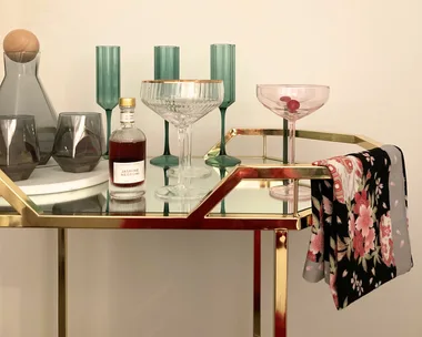 A gold metal and glass bar cart with colourful glassware on top and a floral towel folded over the rail.