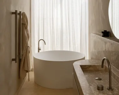 Modern bathroom with a round bathtub, minimalist fixtures, towel rack, and marble countertop beside sheer curtains.