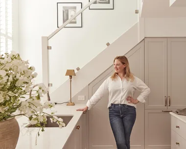 Woman in a white shirt leans against a kitchen counter with flowers in the foreground and a staircase behind her.