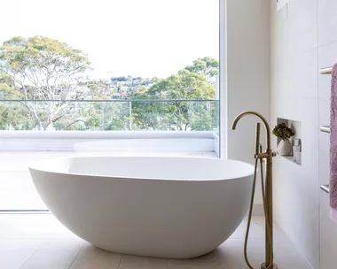 Large pavers cover the floor and wall in this ensuite bathroom, where a large oval-shaped freestanding bath takes centre stage in front of a large window with bushland views.
