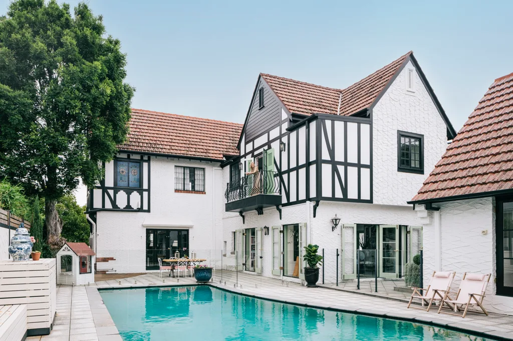 Trending exterior paint colours on a heritage home, including white on the walls and black on the trim. There is a pool beside the Tudor-revival style building.