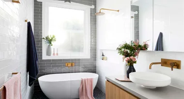 Modern bathroom with freestanding tub, vessel sink, brass fixtures, a vase of flowers, and natural light from a large window.