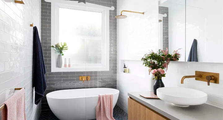 Modern bathroom with freestanding tub, vessel sink, brass fixtures, a vase of flowers, and natural light from a large window.