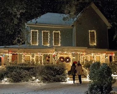 A still from the TV show Gilmore Girls: A Year In The Life. It shows the exterior of Lorelai and Rory's hosue which has been decorated with warm white Christmas lights, with two wreaths on the door. It is snowing.