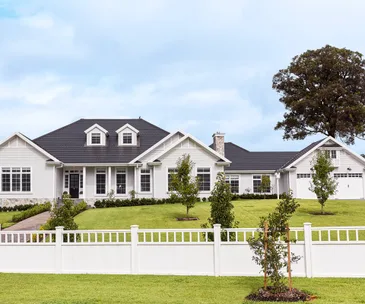 A large white suburban house with a black roof, a spacious green lawn, and a white fence in front.